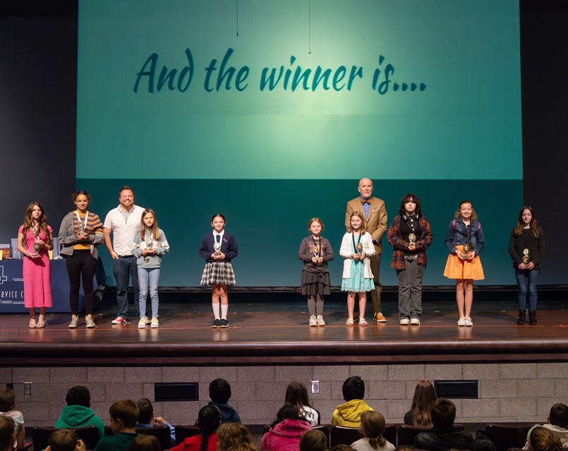 9 GIRLS STANDING IN A ROW AND 2 GUYS BEHIND THEM WAITING TO HEAR WHO WON
